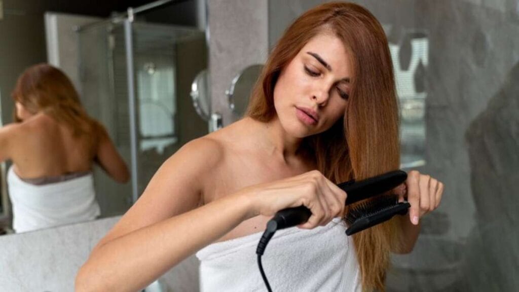 Mulher alisando o cabelo com uma chapinha, diante do espelho do banheiro