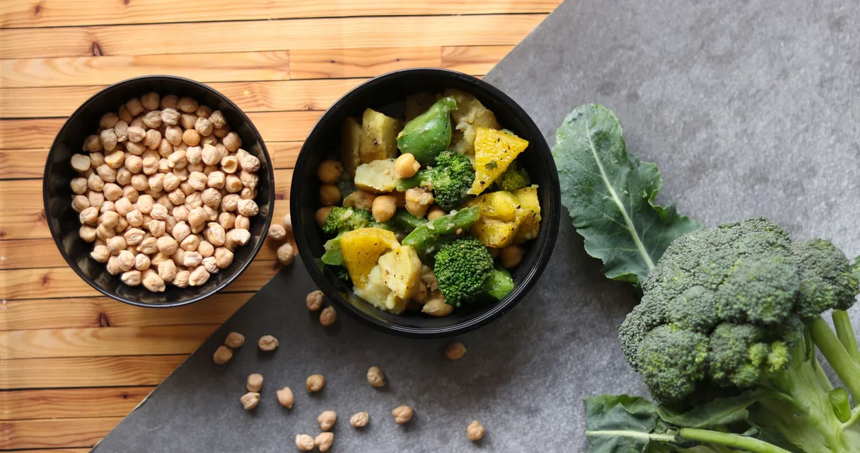 Tigelas pretas com grão-de-bico e legumes cozidos ao lado de um brócolis sobre a mesa