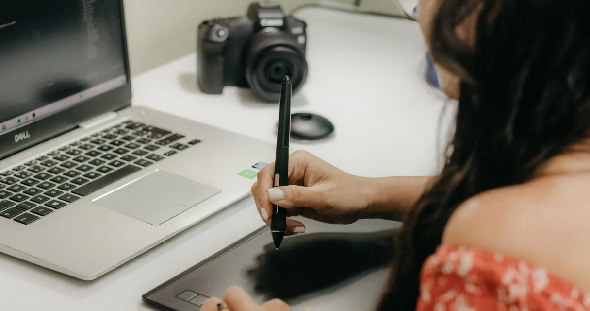 Designer usando mesa digitalizadora e caneta ao lado de um notebook e uma câmera